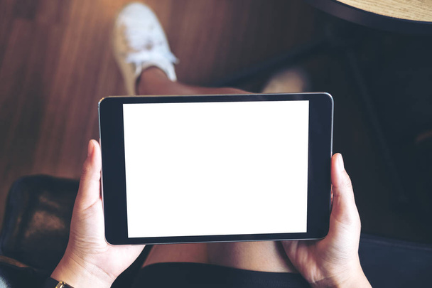Imagen burlona de la mano de la mujer sosteniendo PC tableta negra con pantalla blanca en blanco en el muslo con fondo de madera en la cafetería moderna
 - Foto, imagen
