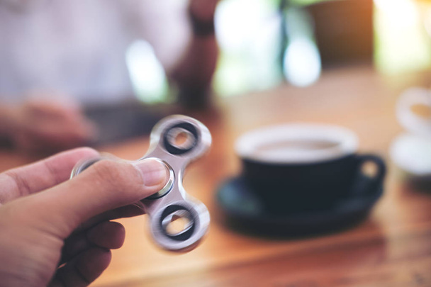 A hand holding and playing metal silver color fidget spinner with coffee cups and people in background - Photo, Image