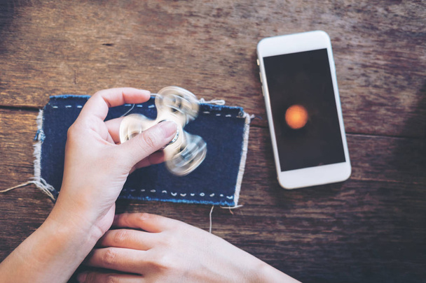 Imagem de vista superior de uma mão segurando e jogando fidget spinner com telefone celular branco com tela preta em branco na mesa de madeira vintage
  - Foto, Imagem