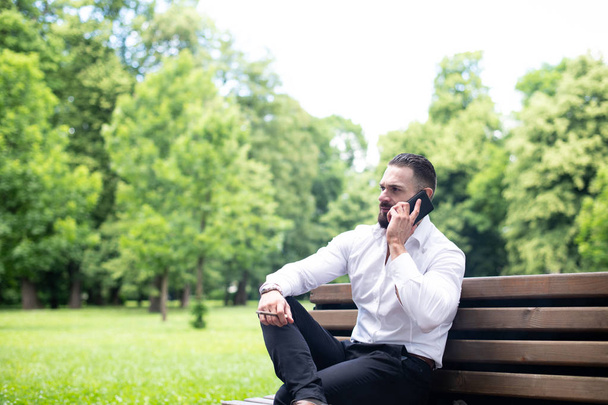 Portrait Of A Young Physically Fit Businessman Smokes Cuban Cigar in Park and Talks on Mobile Phone - Foto, imagen