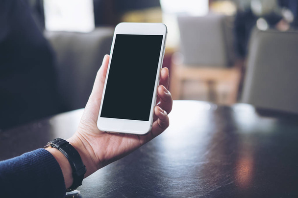 Imagen burlona de la mano de una mujer sosteniendo y mirando el teléfono móvil blanco con pantalla negra en blanco en la oficina moderna
 - Foto, imagen