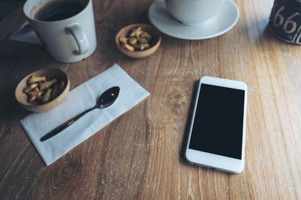 Makieta obrazu biały telefon z puste czarny ekran, filiżanki kawy i przekąski na vintage drewnianym stole w kawiarni - Zdjęcie, obraz