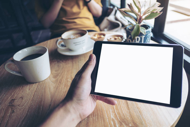 Χλευάζουν την εικόνα ενός χεριού που κρατάει μαύρο tablet pc με λευκές κενές οθόνες στο ξύλινο τραπέζι με γυναίκα που χρησιμοποιούν κινητό τηλέφωνο στο παρασκήνιο στο μοντέρνο καφέ  - Φωτογραφία, εικόνα