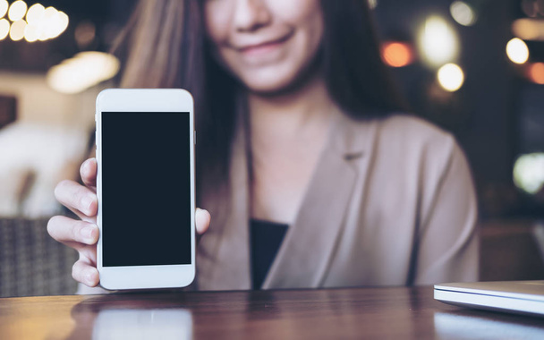 Mockup image of a beautiful Asian business woman holding and showing white mobile phone with blank black screen and smiley face in cafe - Photo, Image