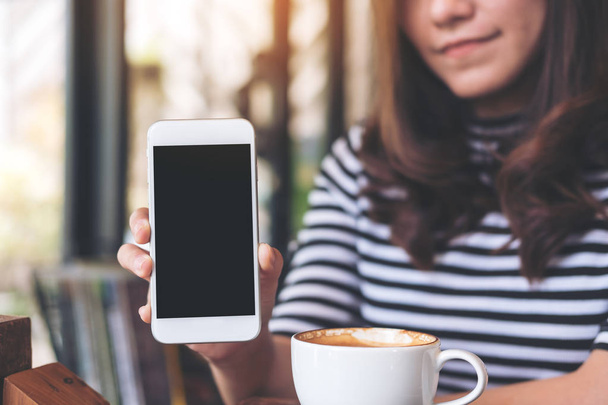 Attrappe einer schönen Frau, die ein weißes Handy mit leerem schwarzen Bildschirm und Kaffeetasse auf dem Tisch im Café hält und zeigt - Foto, Bild