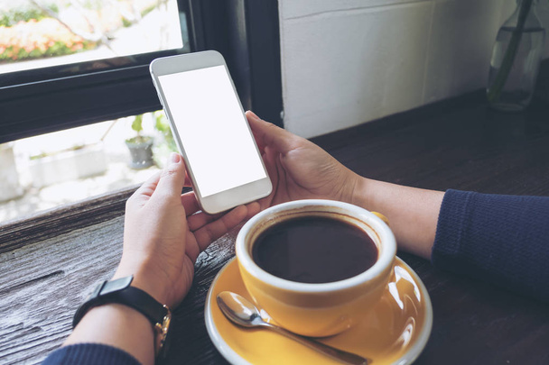 Mockup image of woman's hands holding white mobile phone with blank screen and yellow coffee cup on wooden table in cafe - Photo, Image