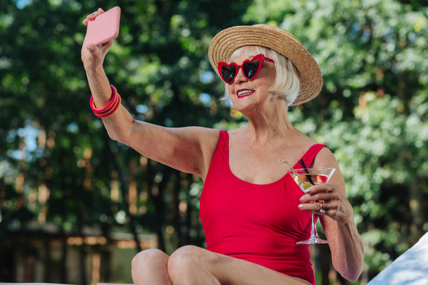 Beaming blonde-haired mature woman making photo with martini - 写真・画像