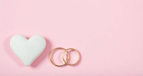 Love and marriage concept. Close up and top view of golden wedding rings and a white heart, isolated, copy space, on a pink background. - Фото, зображення