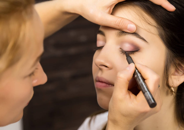 Maquillaje artista aplicando delineador de ojos líquido con cepillo a la cara hermosa mujer. Maquillaje en proceso
 - Foto, Imagen