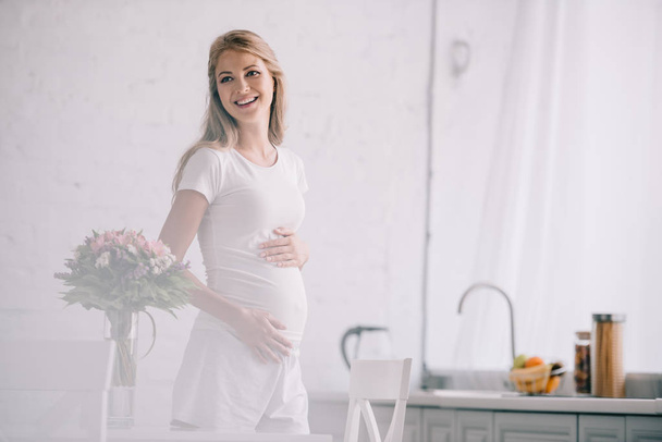 portrait of happy pregnant woman standing at table with bouquet of flowers in vase at home - Photo, Image
