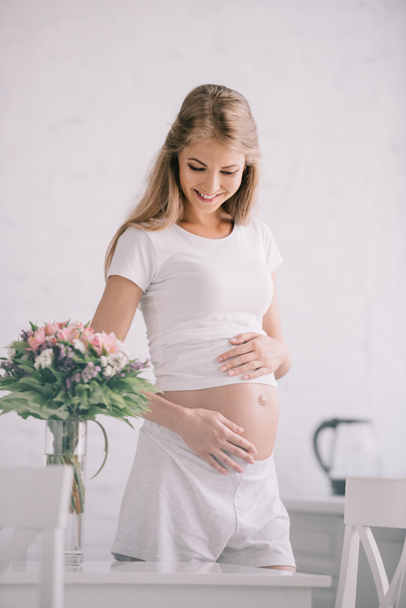 retrato de mujer embarazada feliz de pie a la mesa con ramo de flores en jarrón en casa
 - Foto, Imagen
