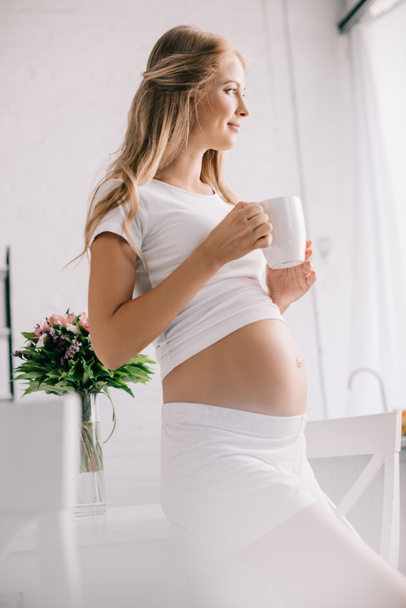 smiling pregnant woman with cup of tea looking away at home - Fotoğraf, Görsel