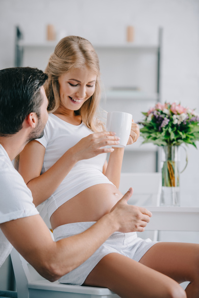 smiling pregnant woman with cup of tea resting on chair with husband near by at home - Photo, Image