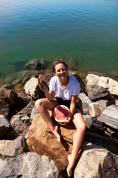 jovem bela mulher rindo alegremente, sorrindo, segurando entre as pernas uma grande melancia e comer, pedras sentadas em um dia de verão perto do mar
 - Foto, Imagem
