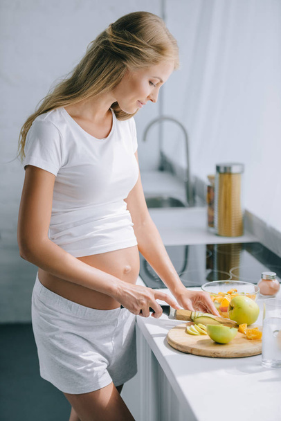 side view of pregnant woman with knife cooking fruits salad at home - Foto, Bild