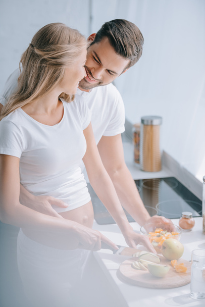 happy pregnant woman and husband cooking fruits salad together in kitchen at home - Photo, image