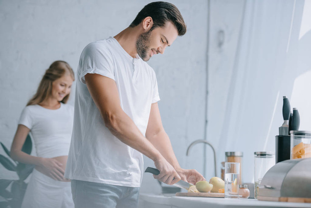 Mann schneidet Obst am Tresen, schwangere Frau in der heimischen Küche - Foto, Bild