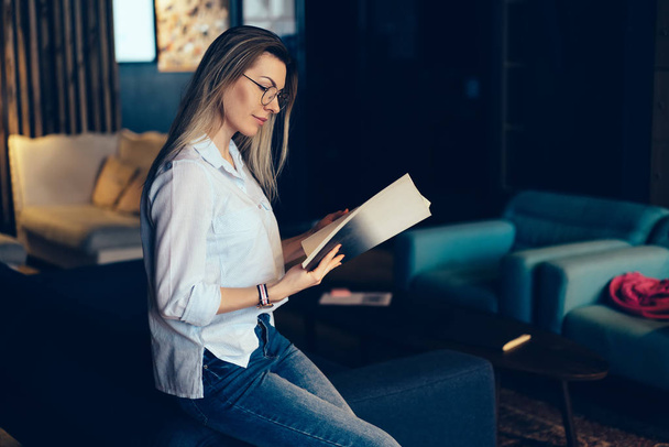 Gorgeous contemplative intelligent woman in glasses for eyes protection reading articles in popular magazine sitting on sofa.Pretty pensive hipster girl enjoying recreation and viewing photos indoors - Valokuva, kuva