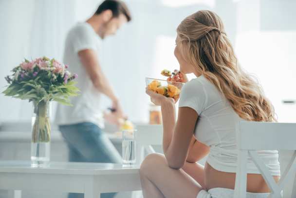 foco seletivo da mulher grávida comendo salada de frutas enquanto o marido de pé no balcão na cozinha
 - Foto, Imagem