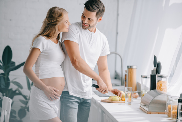 portrét usmívající se muž řezání ovoce na pult s těhotnou manželkou poblíž tím v kuchyni doma - Fotografie, Obrázek