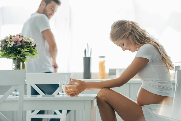 side view of pregnant woman sitting at table with fruits salad in bowl and husband at counter in kitchen - Photo, Image