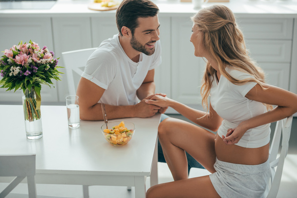happy man and pregnant wife holding hands while sitting at table in kitchen together - Foto, Bild