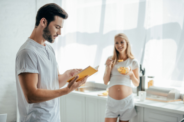 усміхнена вагітна дружина з фруктами салат і чоловік з книгою на кухні вдома
 - Фото, зображення