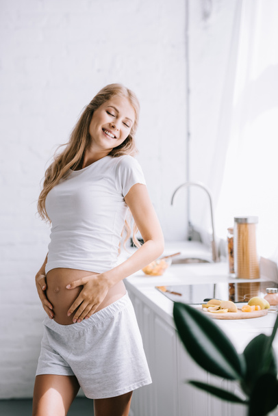 smiling beautiful pregnant woman with hands on belly standing in kitchen at home - Foto, immagini