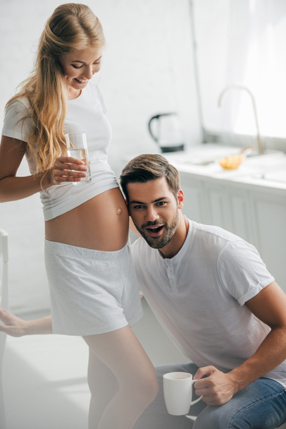 man with cup of coffee listening to belly of pregnant wife in kitchen at home - Foto, Imagen