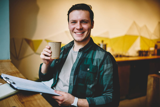 Portrait of young happy male holding folder with article for editing enjoying working day and hot coffee, excited hipster guy satisfied with accountings of successful startup income sitting indoors - Foto, Bild