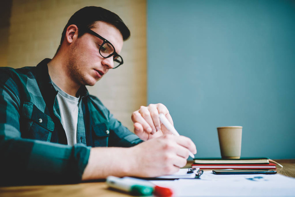 Pensive clever male student looking at wrist watch during writing test quiz in college, concentrated hipster guy checking time organizing learning process preparing for university examination - Foto, Bild