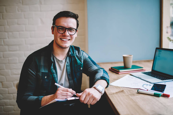 Portrait of cheerful hipster guy in spectacles enjoying studying in coworking space using wifi for online language course, prosperous male freelancer noting idea for publication working remotely - Foto, imagen
