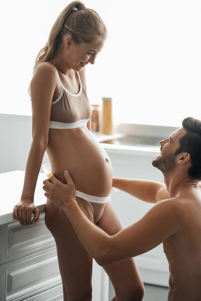vue latérale de l'homme étreignant femme enceinte en sous-vêtements dans la cuisine à la maison
 - Photo, image