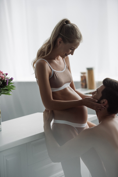 Вид сбоку: мужчина обнимает беременную жену в нижнем белье на кухне дома
 - Фото, изображение