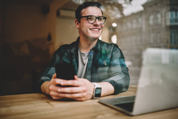 Happy teen guy in spectacles using smartphone for networking sitting in cafe interior with technology,prosperous male freelancer satisfied with working remotely in coffee shop with good wifi - Photo, Image