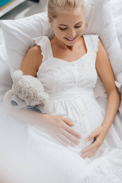 вид сверху на улыбающуюся беременную женщину в белой ночнушке с плюшевым мишкой, лежащим на диване дома
 - Фото, изображение