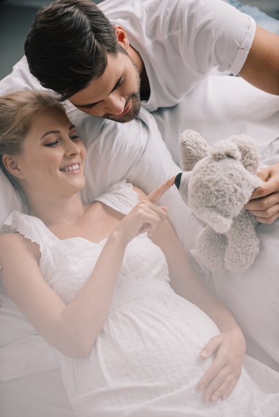 portrait of man with teddy bear and smiling pregnant wife in white nightie on sofa at home - Photo, image