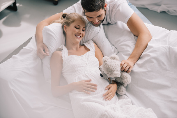 mies nallekarhu lähellä hymyilevä raskaana oleva vaimo valkoinen yöpaita sohvalla kotona
 - Valokuva, kuva
