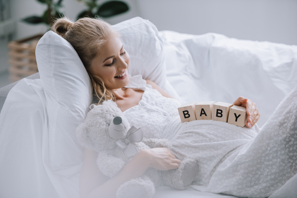 side view of smiling pregnant woman in white nightie with teddy bear and wooden blocks with baby lettering on belly resting on sofa - Photo, Image