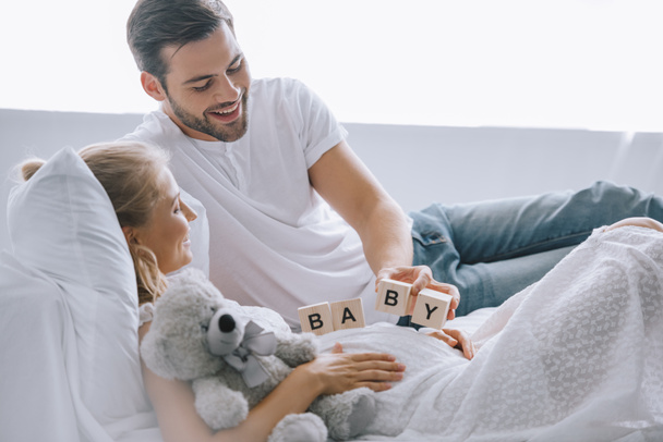 smiling man putting wooden blocks with baby lettering on belly of pregnant wife with teddy bear at home - Photo, Image