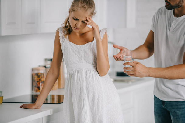 pregnant woman in white nightie having headache while husband giving medicines and glass of water to her at home - Photo, Image