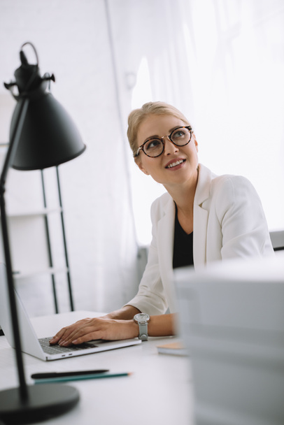 portrait of businesswoman in eyeglasses looking away at workplace with laptop in office - Fotó, kép
