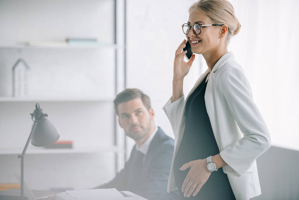 smiling pregnant businesswoman talking on smartphone while standing near colleague at workplace in office - Foto, afbeelding
