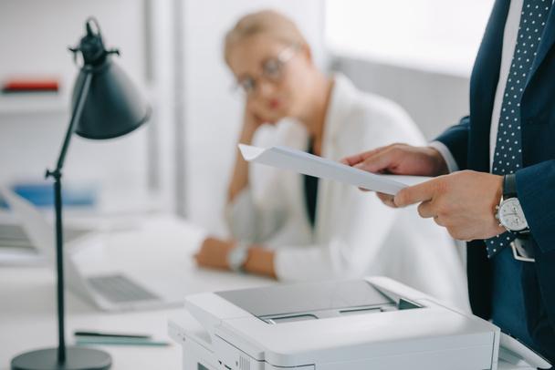selective focus of businessman with papers at printer and businesswoman sitting at workplace in office - Foto, Imagem