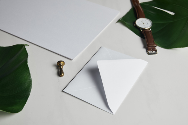 Blank envelope and watch on white marble background with green leaves - Photo, Image