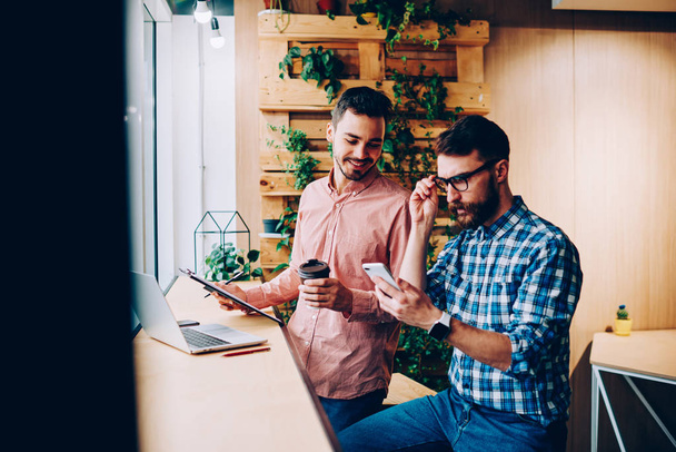 Concentrated hipster guy reading income message on telephone while his friend looking interestly, skilled colleagues making planning on cooperation for creating startup using modern technolog - Photo, image
