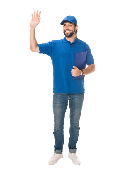 full length view of happy young delivery man holding clipboard and waving hand isolated on white  - Fotoğraf, Görsel