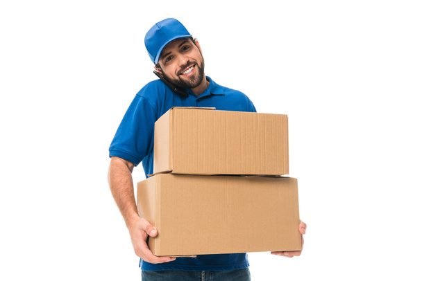 handsome young delivery man holding boxes, talking by smartphone and smiling at camera isolated on white - Φωτογραφία, εικόνα