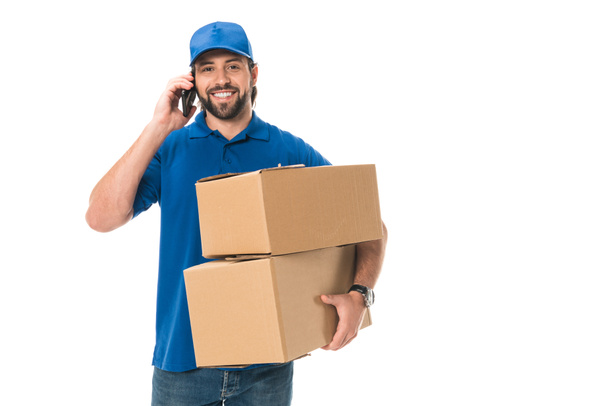 счастливый молодой доставщик держит коробки, разговаривает по смартфону и улыбается на камеру изолированы на белом
 - Фото, изображение