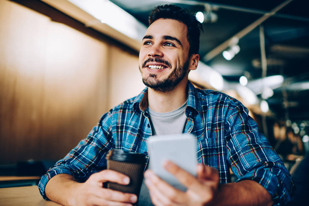 Cheerful handsome guy looking up imagine his vacation while chatting via smartphone in office, young smiling student dreaming about something on coffee break holding mobile phone for networking - Фото, изображение